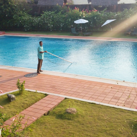 Swimming Pool Cleaning technician using a long-handled brush to clean a large outdoor swimming pool on a sunny day.