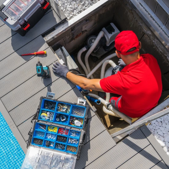 A technician in a red shirt works inside an equipment hatch beside a pool, using tools and a supply box to service pool plumbing.