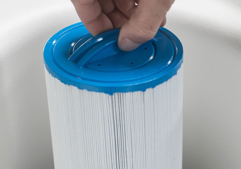 Hand lifting a pleated hot tub filter cartridge by the blue plastic end cap.