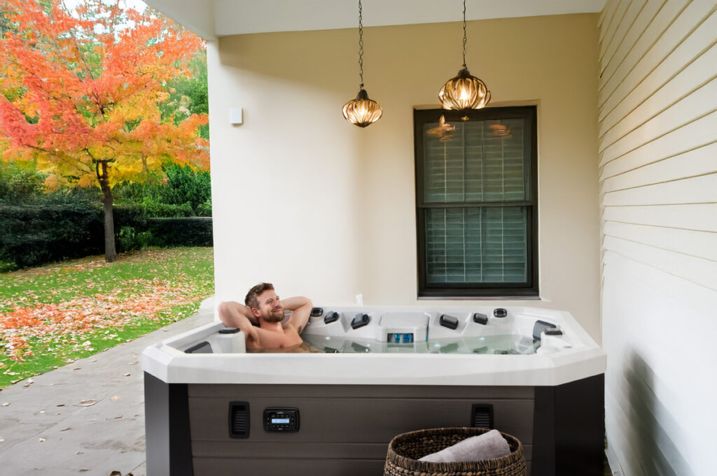 Person relaxing in an outdoor hot tub on a covered porch with autumn trees in the background.